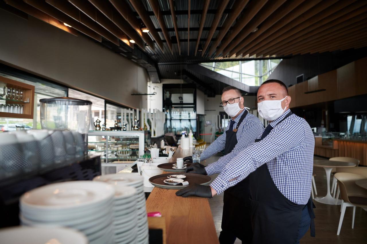 waiter in a medical protective mask serves  the coffee in restaurant durin coronavirus pandemic representing new normal concept.