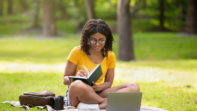 Focused millennial black girl writing something in notebook near laptop computer at park, panorama.