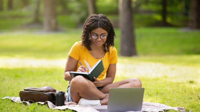 Focused millennial black girl writing something in notebook near laptop computer at park, panorama.