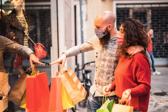 Pretty young man pointing to shop window to show clothing item his likes to his girlfriend - Beautiful young couple enjoying in shopping, having fun together, with the face mask - Consumerism, love, dating, new normal, lifestyle concept.