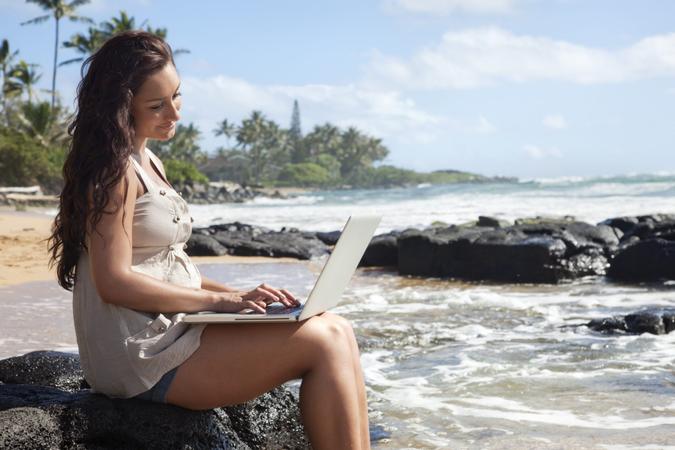 "Photo of a young woman sitting on a remote Hawaiian beach with a laptop, typing.