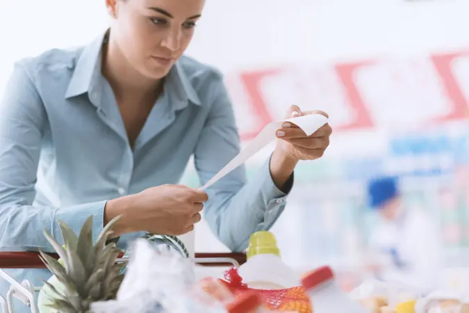 Woman shopping at the supermarket, she is checking a long grocery receipt and leaning on a cart, budgeting and lifestyle concept.