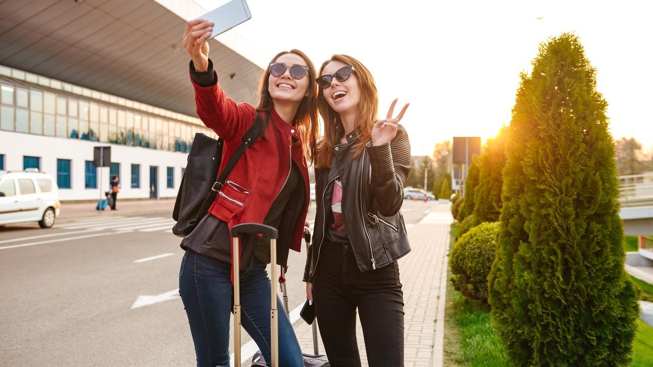 Image of two young caucasian women wearing sunglasses taking selfie photo on smartphone, while standing with luggage near airport waiting for flight or after departure. Air travel concept stock photo