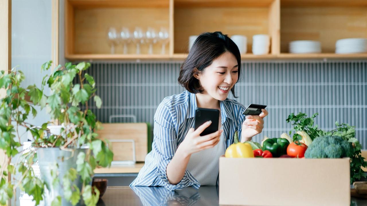 Beautiful smiling young Asian woman grocery shopping online with mobile app device on smartphone and making online payment with her credit card, with a box of colourful and fresh organic groceries on the kitchen counter at home stock photo