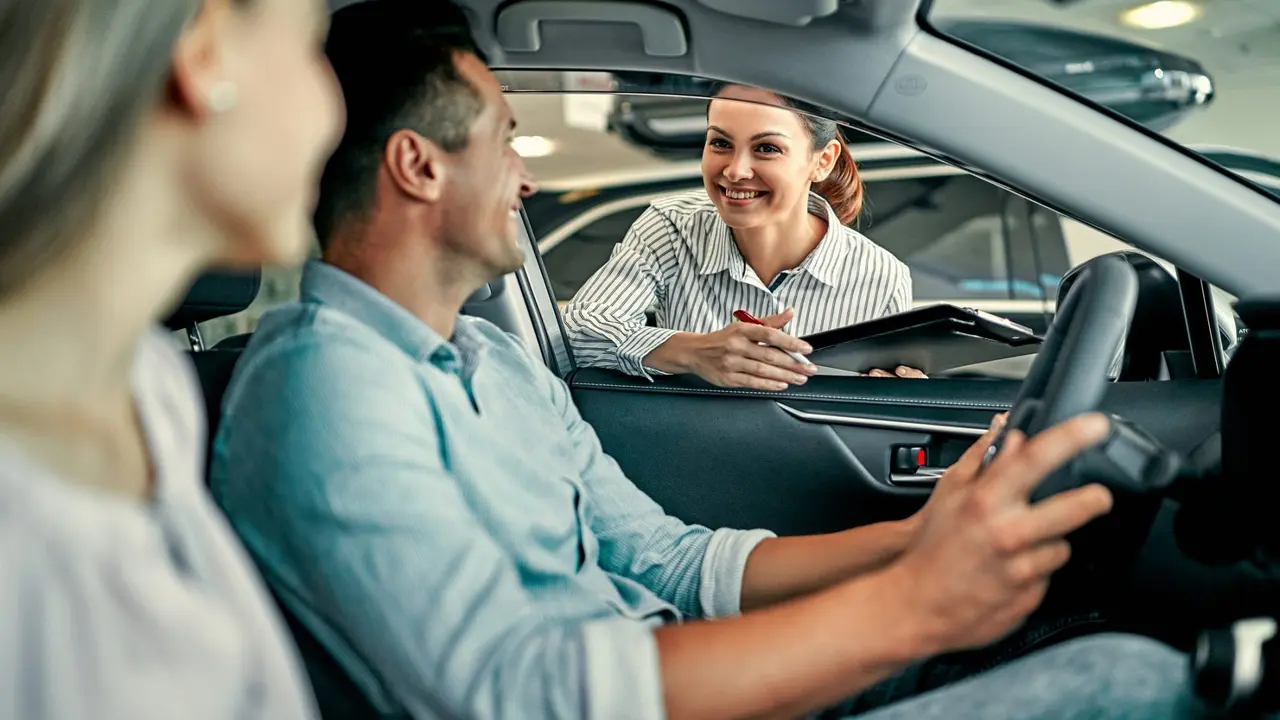 Young female consultant and couple buyers sitting in new car signing contract in auto show.