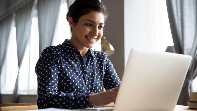 Smiling indian girl student professional employee typing on laptop sit at home office table, happy hindu woman studying e learning online software using technology app for work education concept.