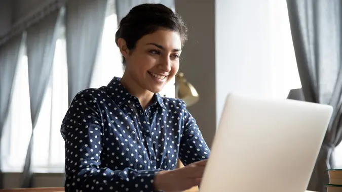 Smiling indian girl student professional employee typing on laptop sit at home office table, happy hindu woman studying e learning online software using technology app for work education concept.