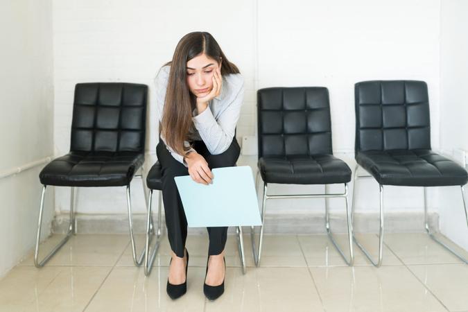 Bored Hispanic female applicant sitting with resume in waiting room during recruitment at office.