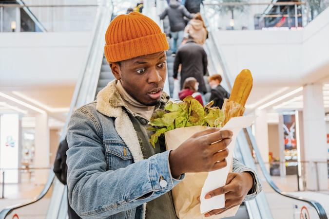 African man with a paper bag of groceries looks surprised and upset at a receipt from a supermarket with high prices against the background of an escalator with customers in the shopping center.