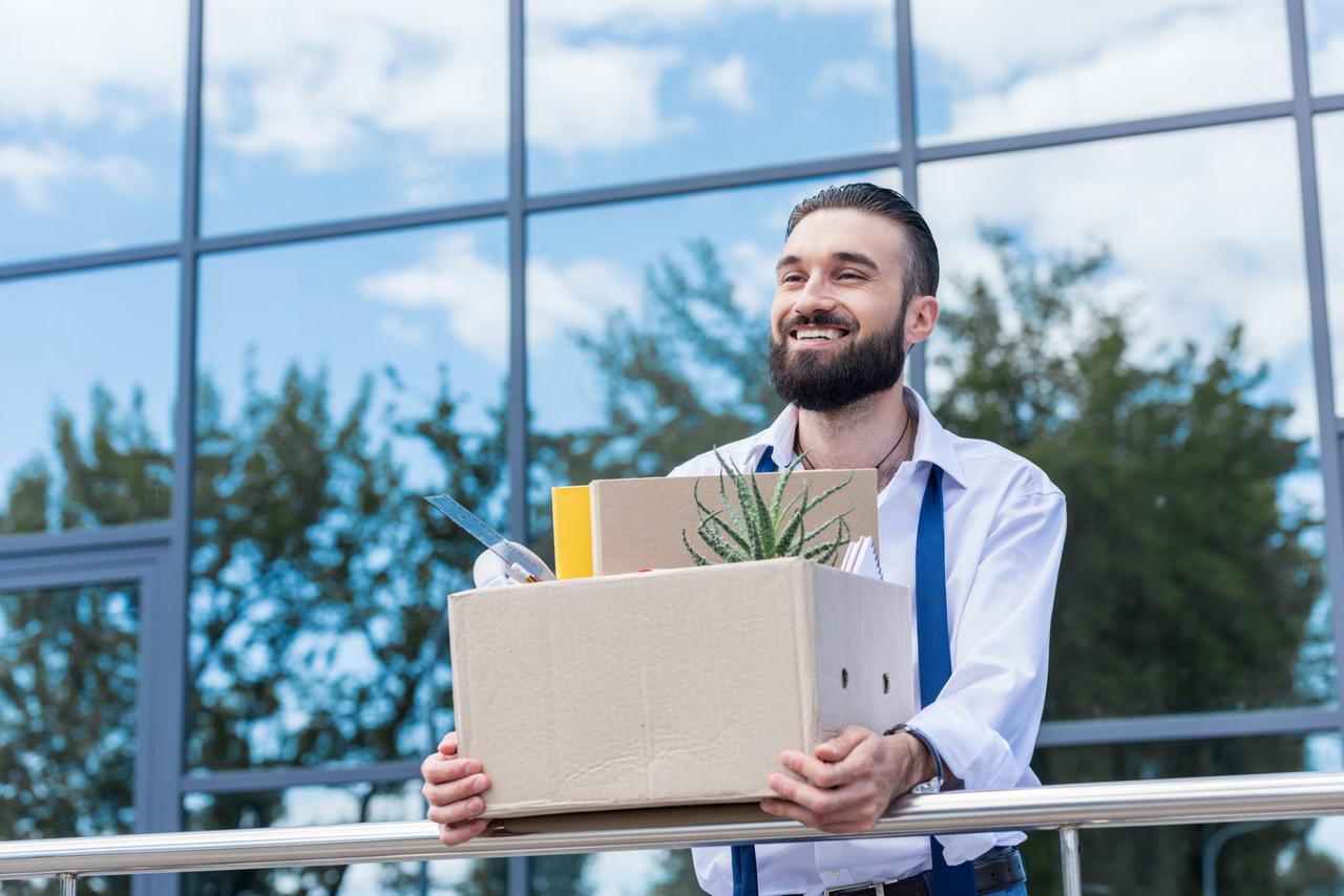 happy businessman with cardboard box with office supplies in hands standing outside office building, quitting job concept.