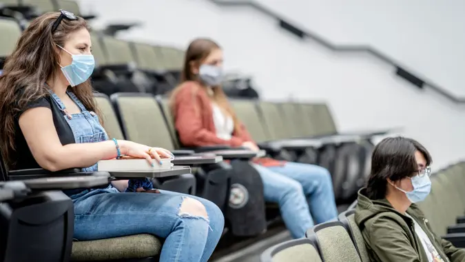 Multi-ethnic group of students wearing protective face masks while sitting in a lecture hall sitting 2 meters apart.