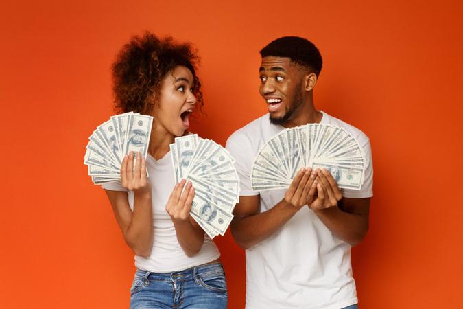 Excited african american couple holding bunch of money banknotes and looking at each other in amazement, orange studio background.