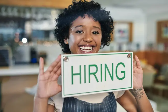 Portrait of a young woman holding a "hiring" sign in her store.