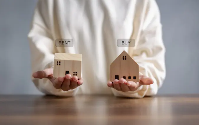 Hand holding wooden home with text message Buy or Rent, on wooden desk office.
