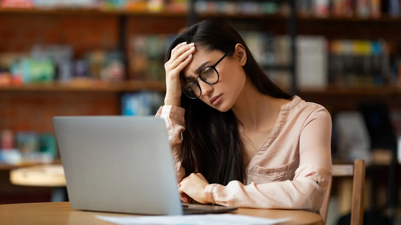 Woman using a laptop in a cafe, resting her head on one hand and looking unhappy.