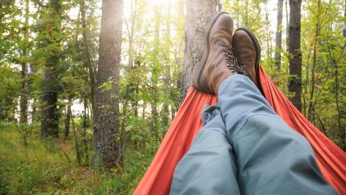 Person wearing trekking pants and hiking boots relaxing laying in camping hammock overlooking summer forest at sunset.