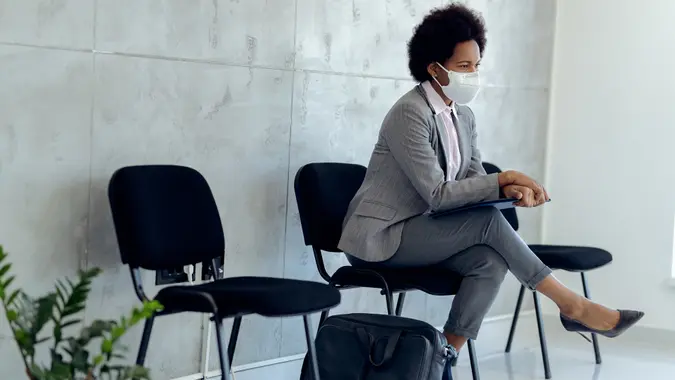 Distraught African American businesswoman wearing protective face mask while sitting in a hallway and waiting for job interview.