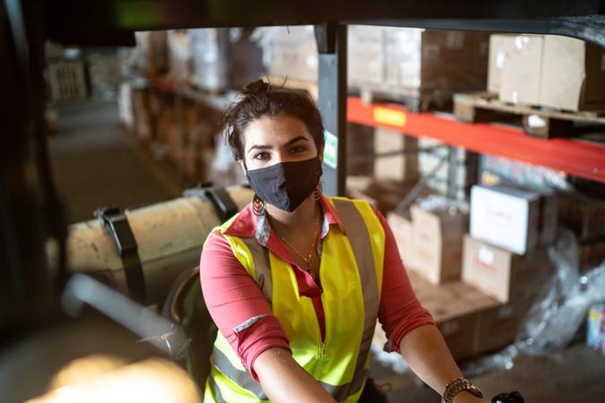Portrait of a young woman using face mask driving a forklift in a warehouse.
