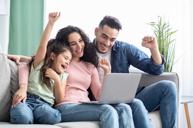 Portraif of happy arabic family of three celebrating success with laptop at home, cheerful middle eastern mother, father and daughter looking at computer screen and raising hands with excitement.