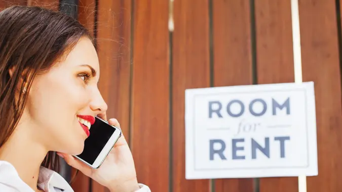 caucasian woman in white shirt standing in front of wooden fencing with "room for rent" ads and calling to house owner.