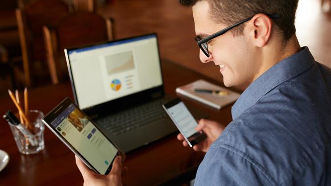 Attractive man in glasses working with multiple internet electronic devices.  Freelance businessman has laptop and smartphone in hands and laptop on table with charts on screen.  Multitasking theme stock photo