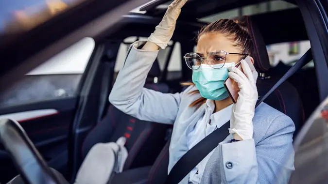Businesswoman with protective mask and gloves on sitting in her car caught in traffic jam sitting in her car and having phone conversation. She is very nervous. Protection form corona virus concept. stock photo