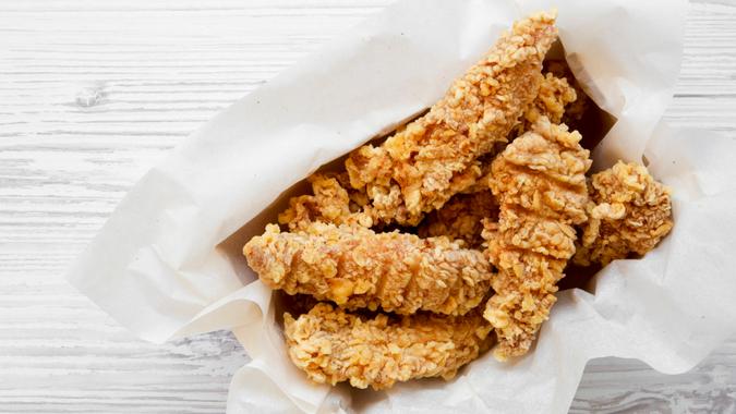 Chicken strips in paper box on a white wooden table, top view.