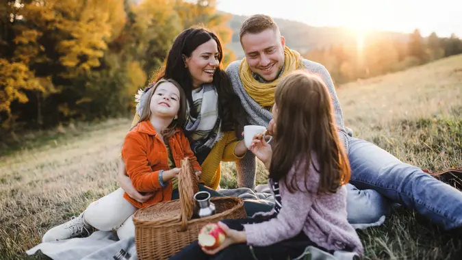 A portrait of happy young family with two small children sitting on a ground in autumn nature at sunset, having picnic.