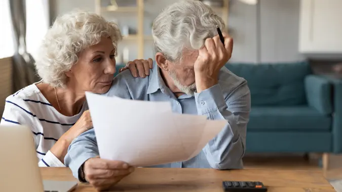 Distressed mature 60s husband and wife sit at table calculate manage household expenses having problems with finances, upset pensioners frustrated troubled with paying bills expenditures.