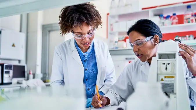 Two female scientists talking together and writing data in a notebook while working at a table in a lab.