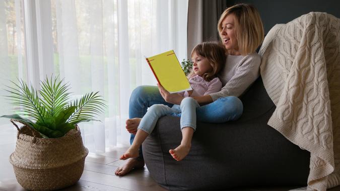 Mother reading book to her little daughter, sitting on a bean bag chair at home.