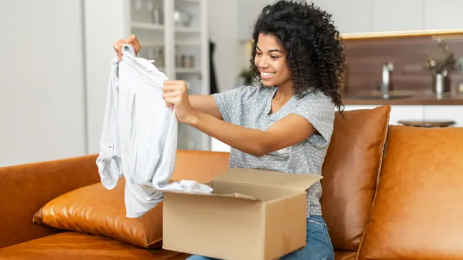 Satisfied smiling young African American woman sitting on the sofa opening parcel carton box, unpacking received gift, item from an online store, female customer happy with fast delivery and shipment.