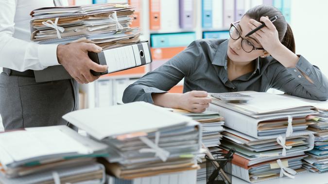 Young stressed secretary in the office overwhelmed by work and desk full of files, her boss is bringing more paperwork to her.