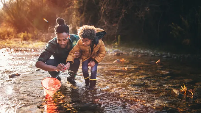 Young dad teaching son how to fish with fishing net in mountain stream at sunset.