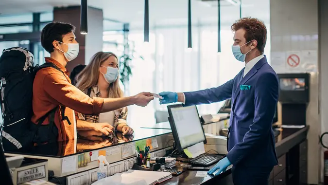 Three people, young couple travelers with medical masks on hotel reception talking to male receptionist.