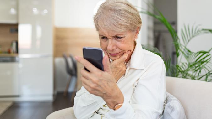 Confused senior woman having trouble using mobile phone at home. Old woman with white hair sitting on sofa and trying to messaging with smartphone. stock photo