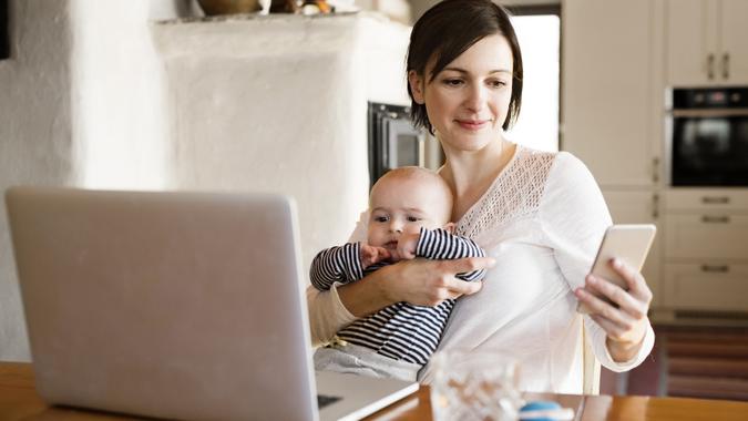 Mother at home with baby working on laptop, holding smartphone stock photo