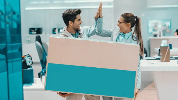 Smiling happy multicultural couple dressed elegant holding box with new plasma tv and giving high five to each other while standing in tech store.