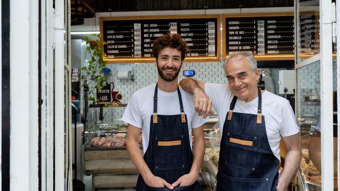 Father and so owners of a butcher shop standing at the entrance smiling at camera very happy.