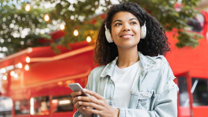 African-american young woman student freelancer teenager listening to the music in headphones earphones choosing sound track song singer playlist on smart phone outdoors in park.