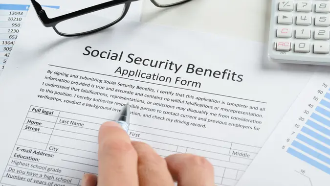 Close-up Of Hand With Pen And Eyeglasses Over Social Security Benefits Application Form.
