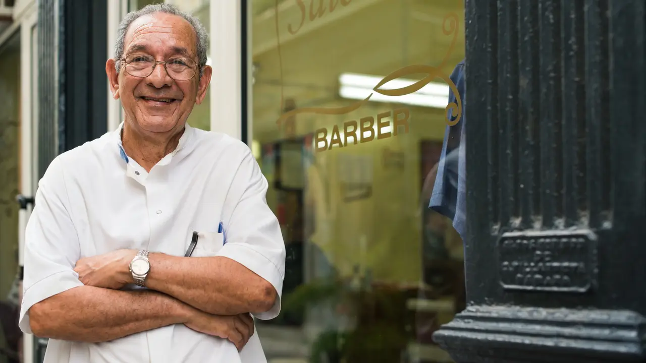 senior hispanic barber in old fashion barber's shop, posing and looking at camera with arms crossed near shop window.