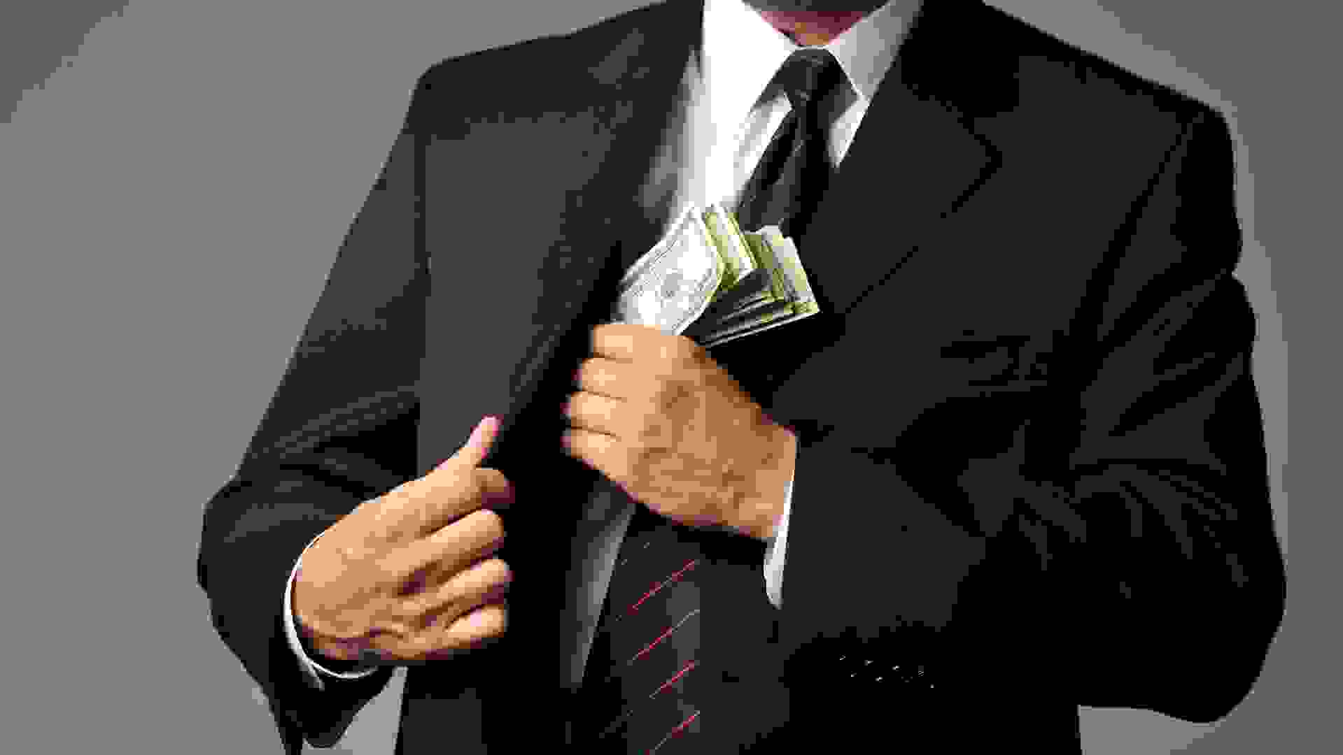 Horizontal studio image of a well dressed businessman putting a stack of $100 bills in his suit coat pocket.