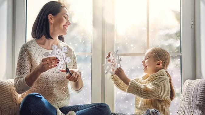 Merry Christmas and happy holidays! Happy loving family sitting by the window and making paper snowflakes for decoration windows.