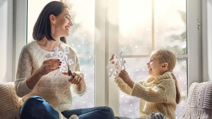Merry Christmas and happy holidays! Happy loving family sitting by the window and making paper snowflakes for decoration windows.