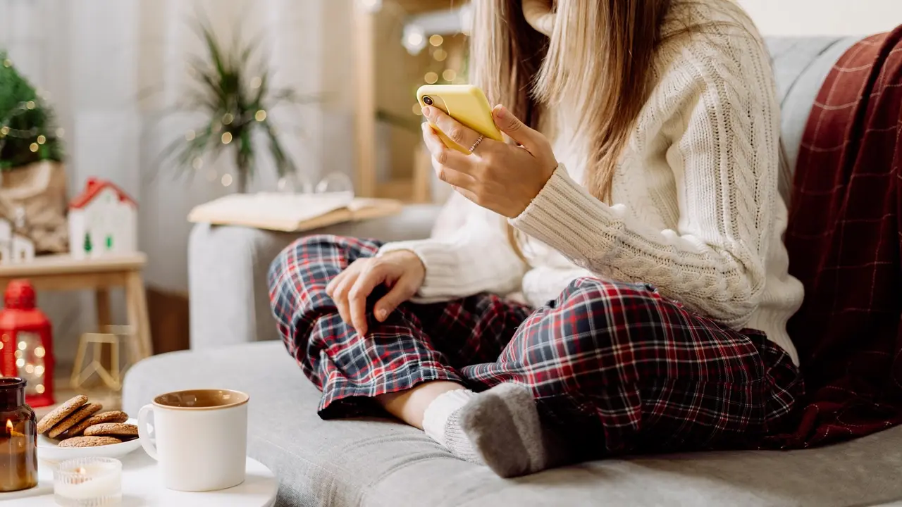 Cozy woman in knitted winter warm socks, sweater and checkered plaid with phone, drinking hot cocoa or coffee in mug, during resting on couch at home. Christmas holidays with candle, decor and cookie stock photo