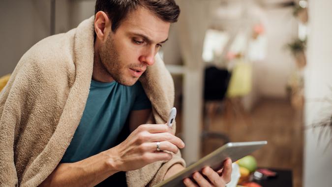 Ill young man sitting at home under blanket and holding digital tablet in one hand and thermometer in other.