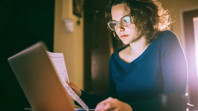 Serious girl studying and working, using laptop and papers, sitting in a living room.