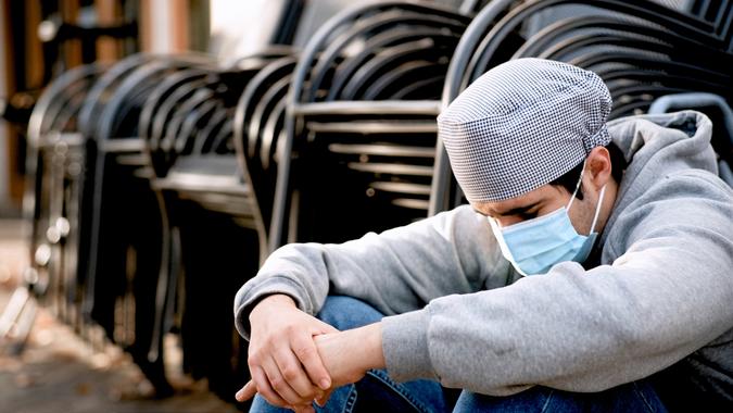 Serious pensive Young cook posing wearing protective face mask sitting on the floor outside the restaurant he works that its closed because the pandemic state at home order.