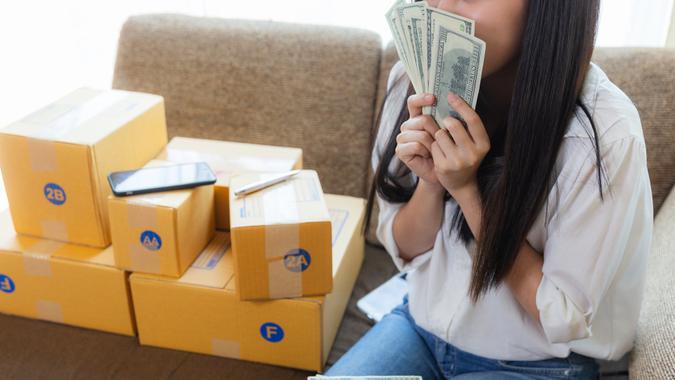 Une jeune femme asiatique démarre des propriétaires de petites entreprises avec une boîte pour la livraison en ligne au client, une jolie fille tenant de l'argent en dollars américains beaucoup d'argent un sourire heureux travaille à la maison.